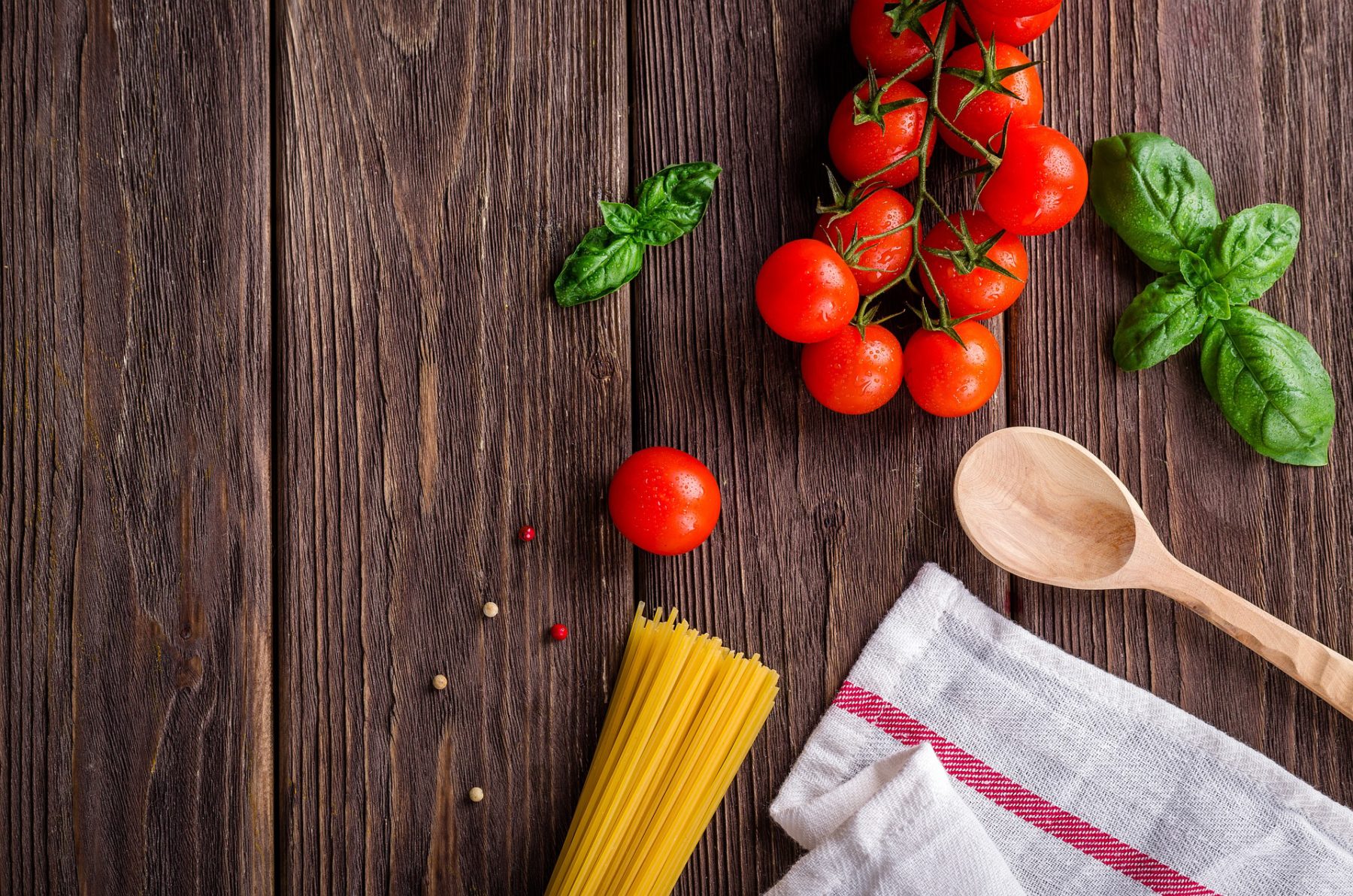 dark stained wood background with cherry tomatoes, uncooked spaghetti noodles, a white towel with a red stripe, a wooden spoon and basil leaves on it