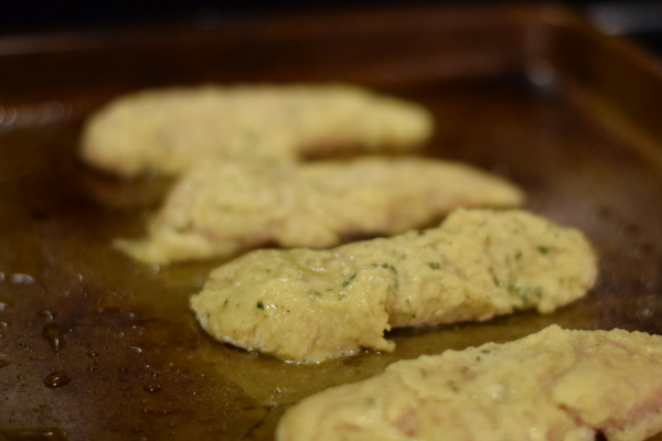 4 raw chicken tenders breaded and ready to be cooked on a dark baking sheet